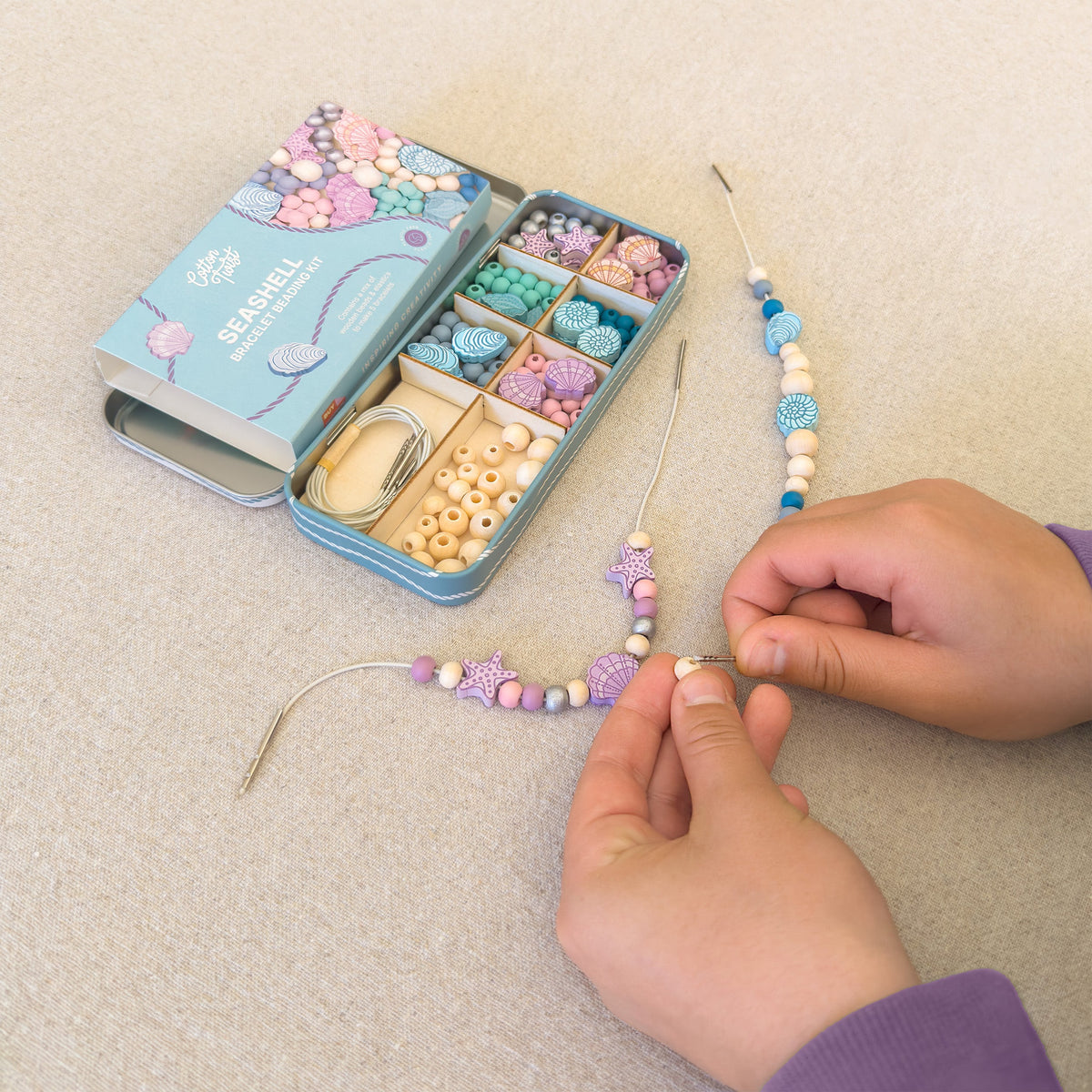 Hands threading seashell wooden beads from the Seashell Beading Kit tin