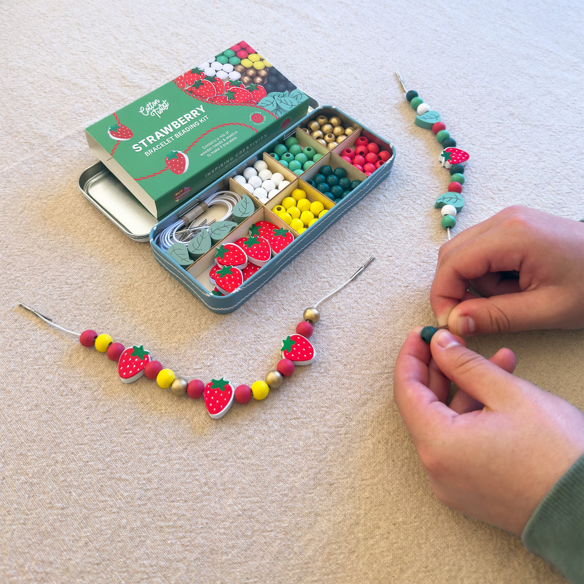 Hands threading strawberry wooden beads from the Strawberry Beading Kit tin