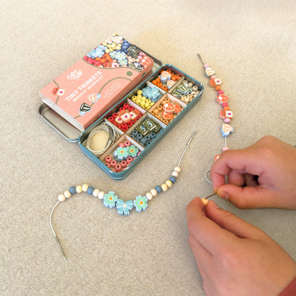 Hands threading wooden beads from the Tiny Trinkets Beading Kit tin